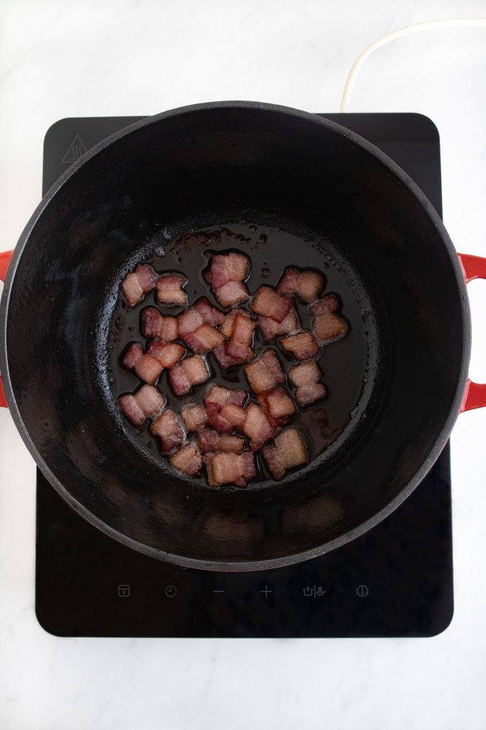 Crispy pieces of bacon are cooking in a black cast iron pot on an electric stovetop with a white countertop background.