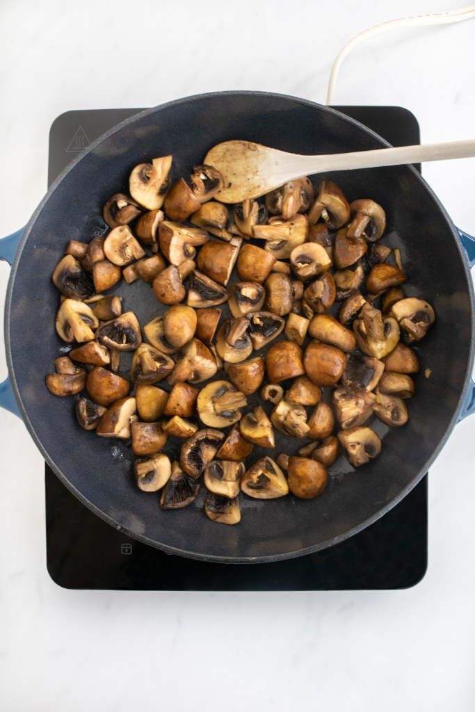 A blue skillet on a stovetop contains sautéed, sliced brown mushrooms being stirred with a wooden spoon. The stove rests on a white countertop.