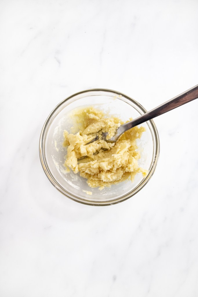A glass bowl containing a mixture of dough or batter being stirred with a metal fork, placed on a white marble surface.
