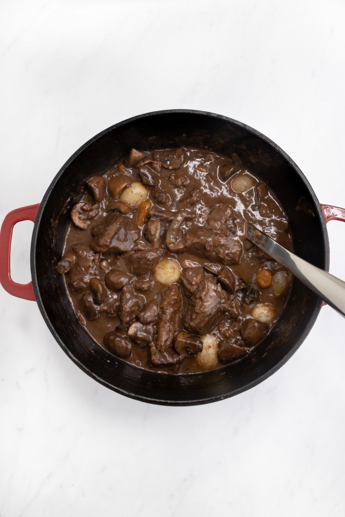 A red pot filled with beef stew, featuring chunks of beef, mushrooms, and pearl onions in a rich brown sauce, with a serving spoon resting inside. The pot sits on a white marble surface.