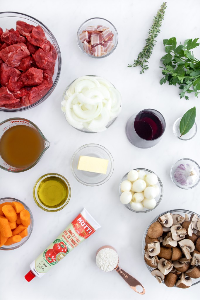 Top-down view of assorted ingredients for a stew, including raw beef, bacon, carrots, mushrooms, onions, pearl onions, garlic, herbs, butter, olive oil, tomato paste, flour, beef broth, and red wine on a white surface.