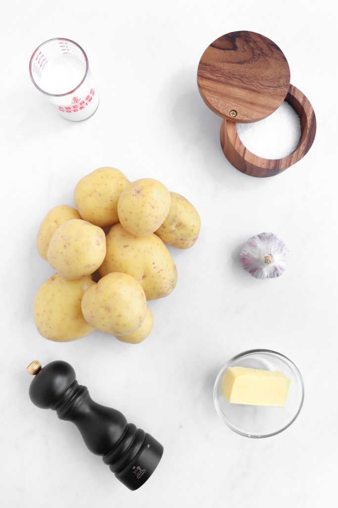 A pile of yellow potatoes, a garlic bulb, a glass of milk, a small glass bowl of butter, a wooden salt cellar, and a black pepper grinder arranged on a white surface.
