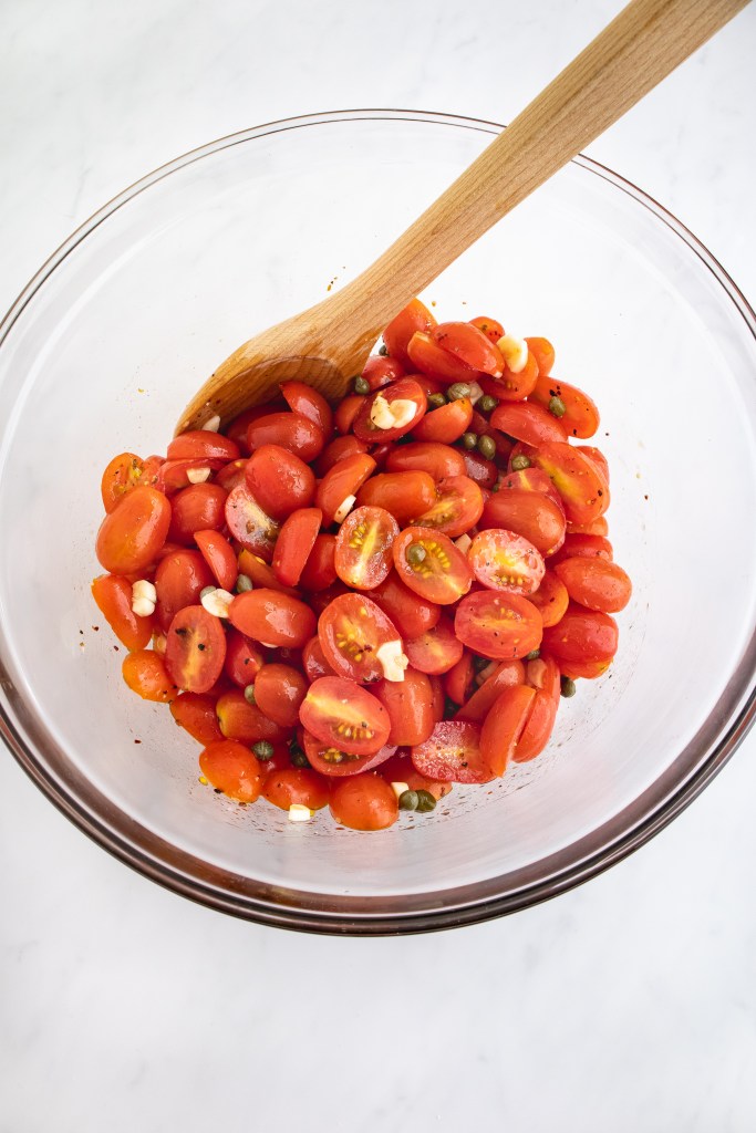 A glass bowl filled with halved cherry tomatoes, capers, and pieces of garlic, mixed with a wooden spoon on a white marble surface.