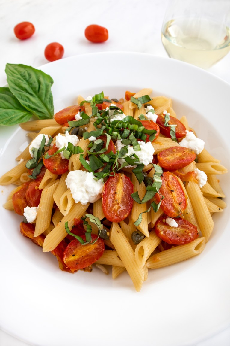 A white plate of penne pasta topped with roasted cherry tomatoes, dollops of goat cheese, and fresh chopped basil leaves. A sprig of basil is on the side, with cherry tomatoes and a glass of white wine in the background.