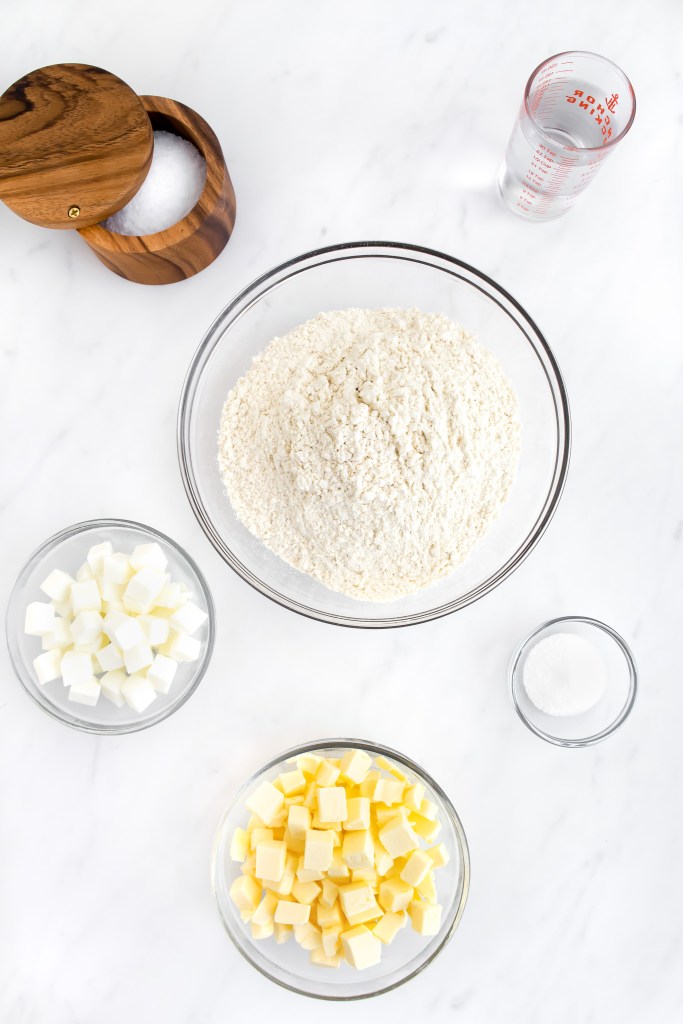 Ingredients for flaky pie crust with salt, water, flour, salt, vegetable shortening and butter on a marble countertop