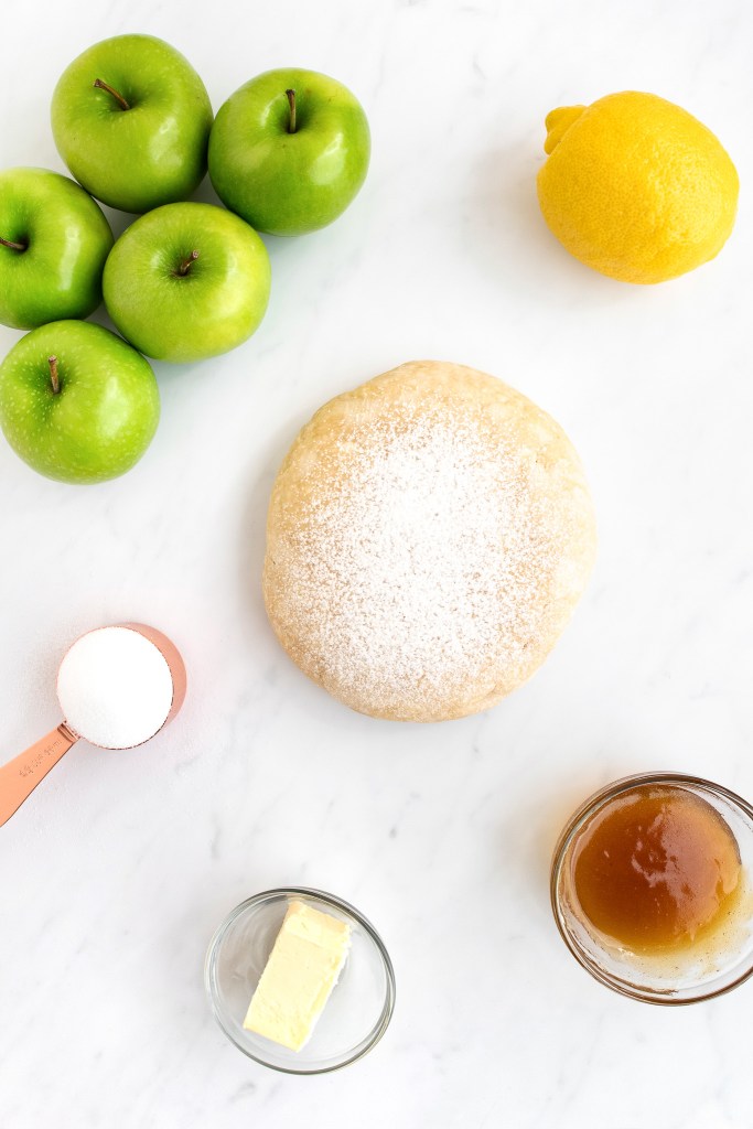 Ingredients for French apple tart with Granny Smith apples, a lemon, pastry dough, sugar, butter and apricot glaze on marble countertop