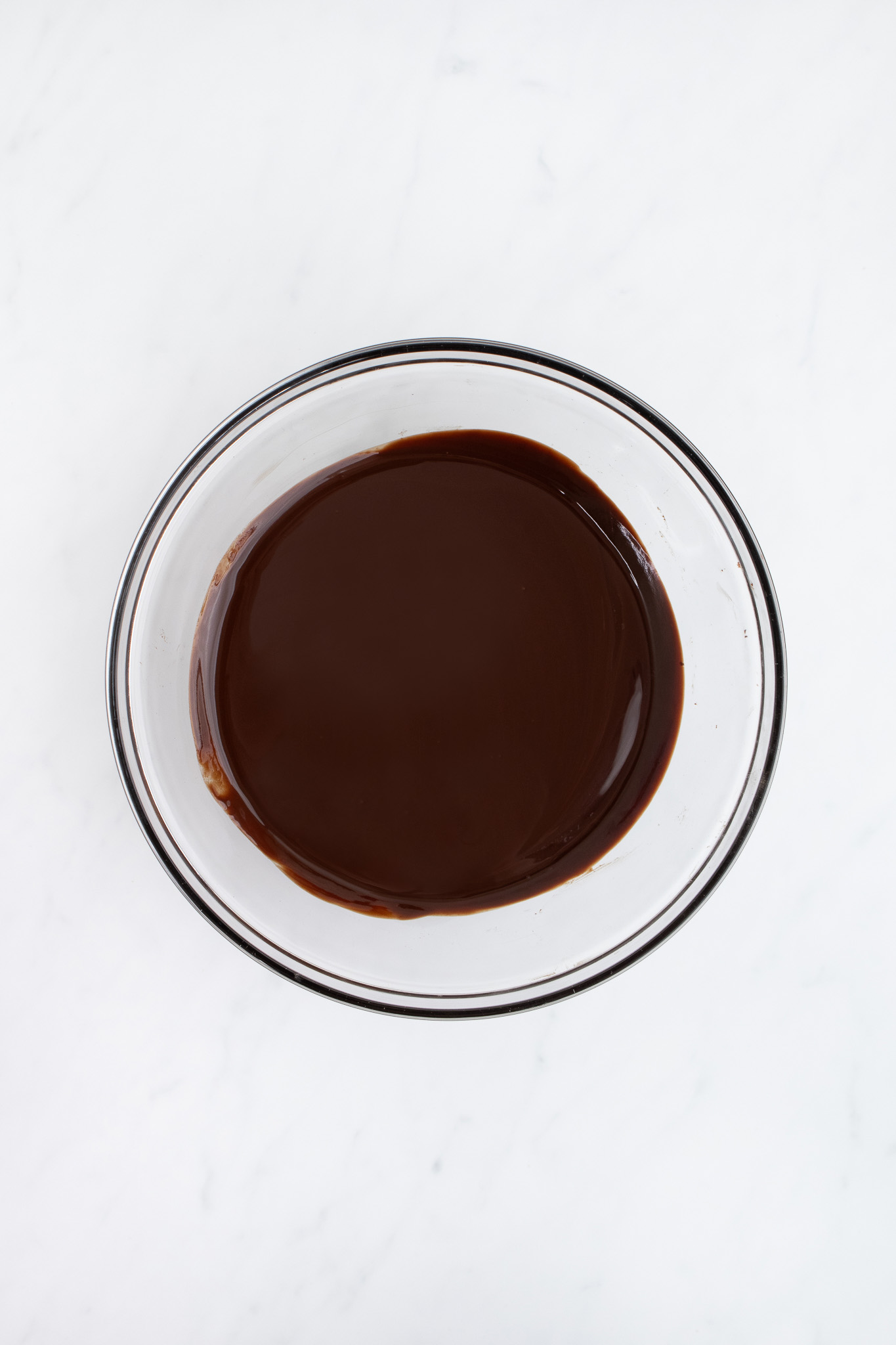 A clear glass bowl filled with smooth, melted chocolate for chocolate mousse sits on a white marble surface.