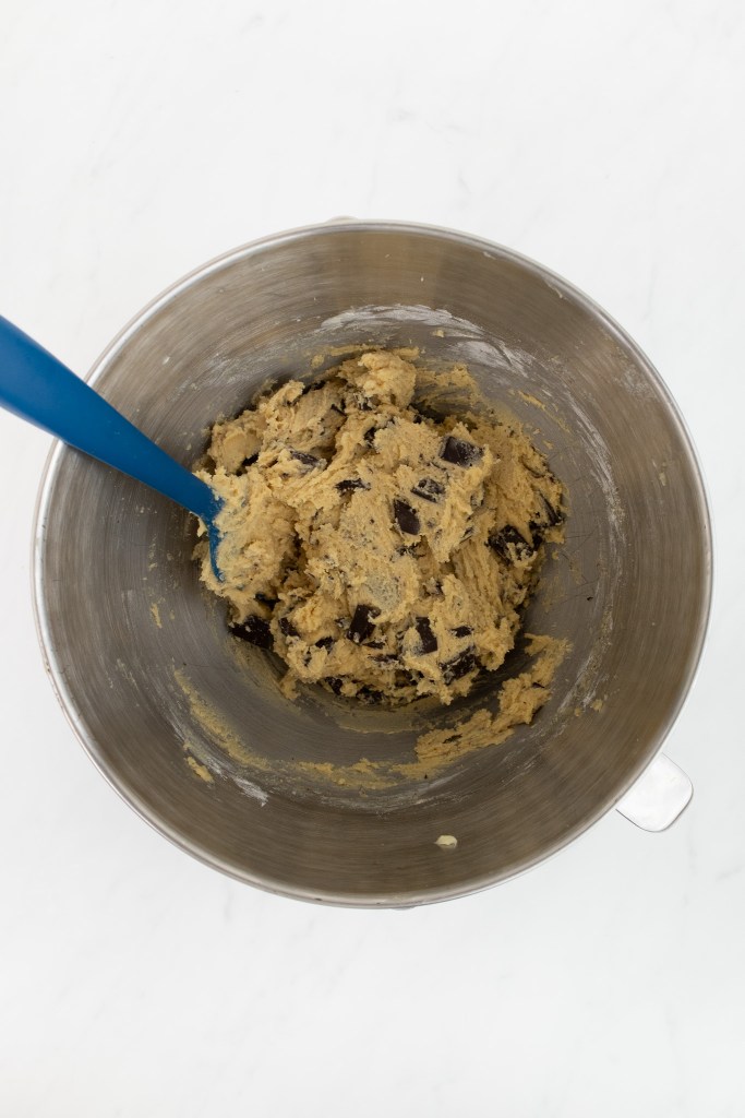 The bowl of an electric stand mixer with chocolate chip cookie dough and a blue spatula, resting on a white marble surface.