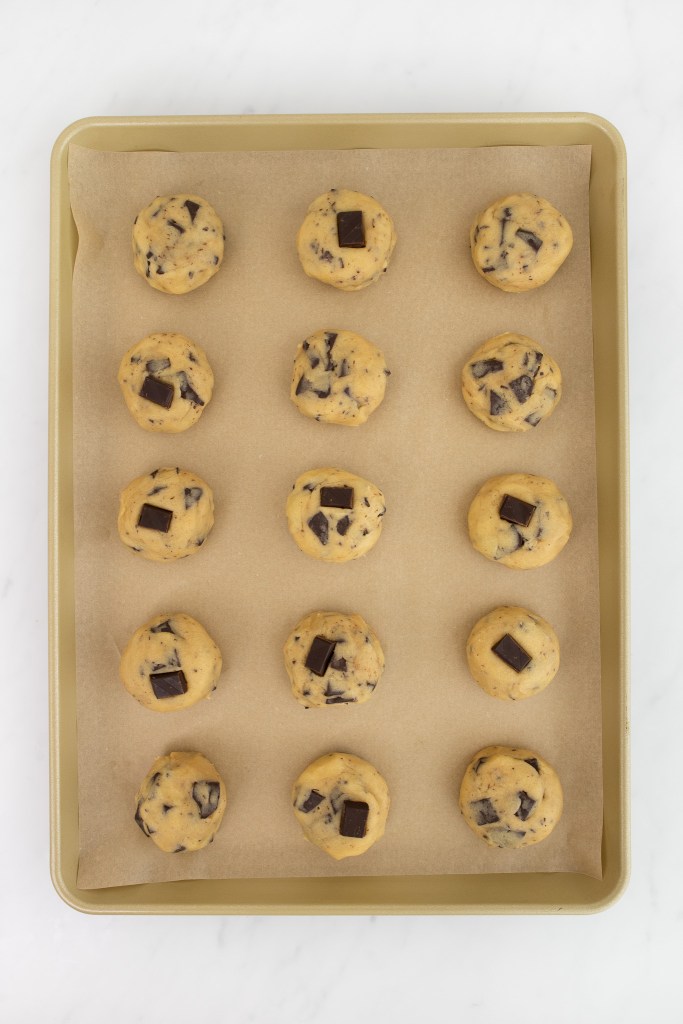 A baking tray lined with parchment paper holds fifteen evenly spaced balls of chocolate chip cookie dough.
