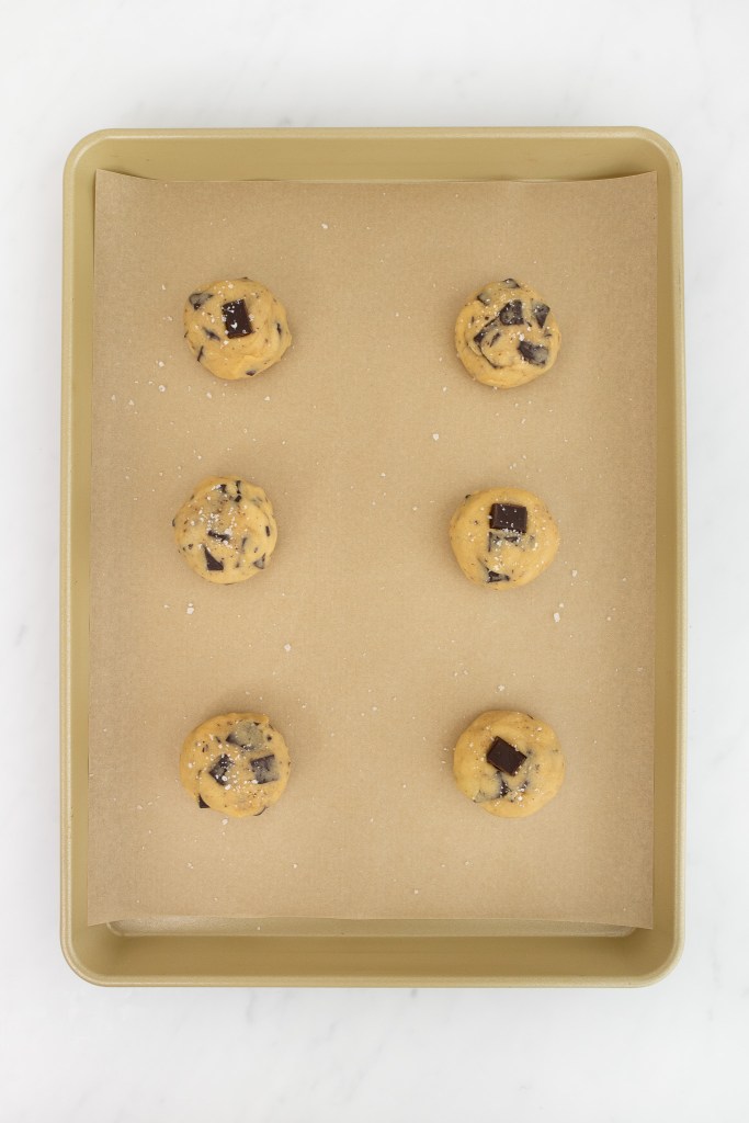 A baking sheet lined with parchment paper holds six unbaked chocolate chip cookie dough balls, sprinkled with coarse salt and ready for the oven.