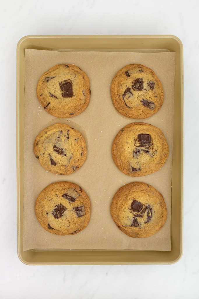 A baking tray lined with parchment paper holds six baked chocolate chip cookies arranged in two columns of three on a white marble surface.