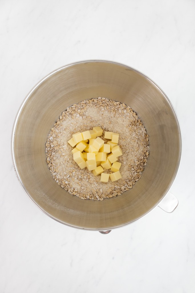 The bowl of an electric stand mixer contains oat mixture with unmixed cubes of butter, viewed from above on a white marble surface.