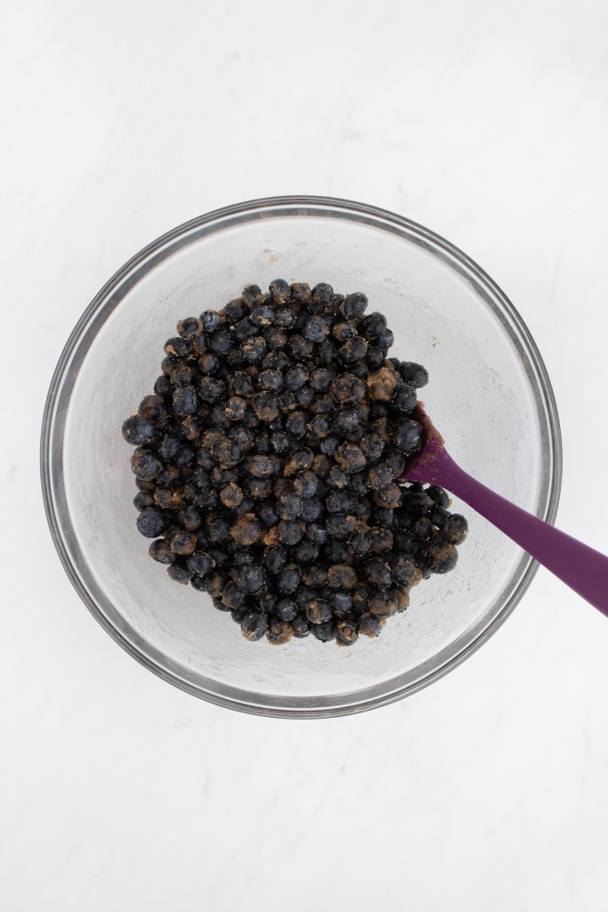 A clear glass bowl filled with blueberries coated in a sugar and cornstarch mixture, with a purple spatula resting inside, on a white marble surface.