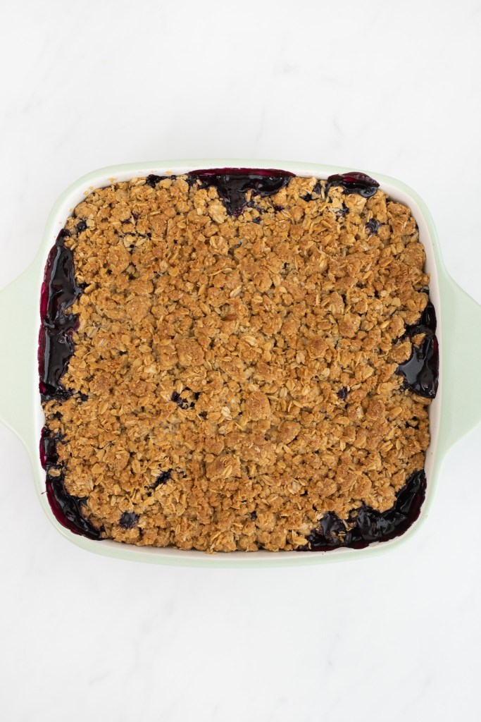 A square baking dish filled with fully baked blueberry crisp sits on a white marble surface.