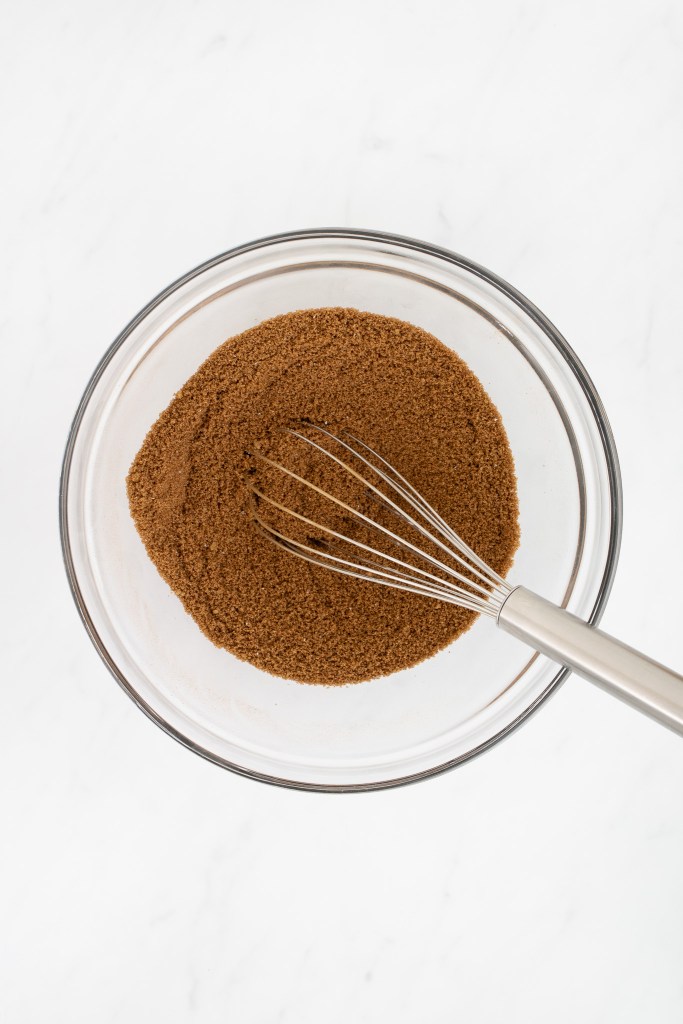 A metal whisk rests in a glass bowl filled with brown cinnamon sugar on a white marble surface.