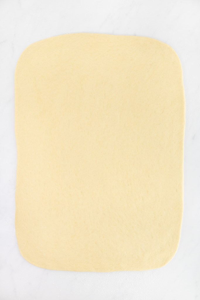 A large rectangle of dough is rolled out on a white marble surface, ready to be used for cinnamon rolls.