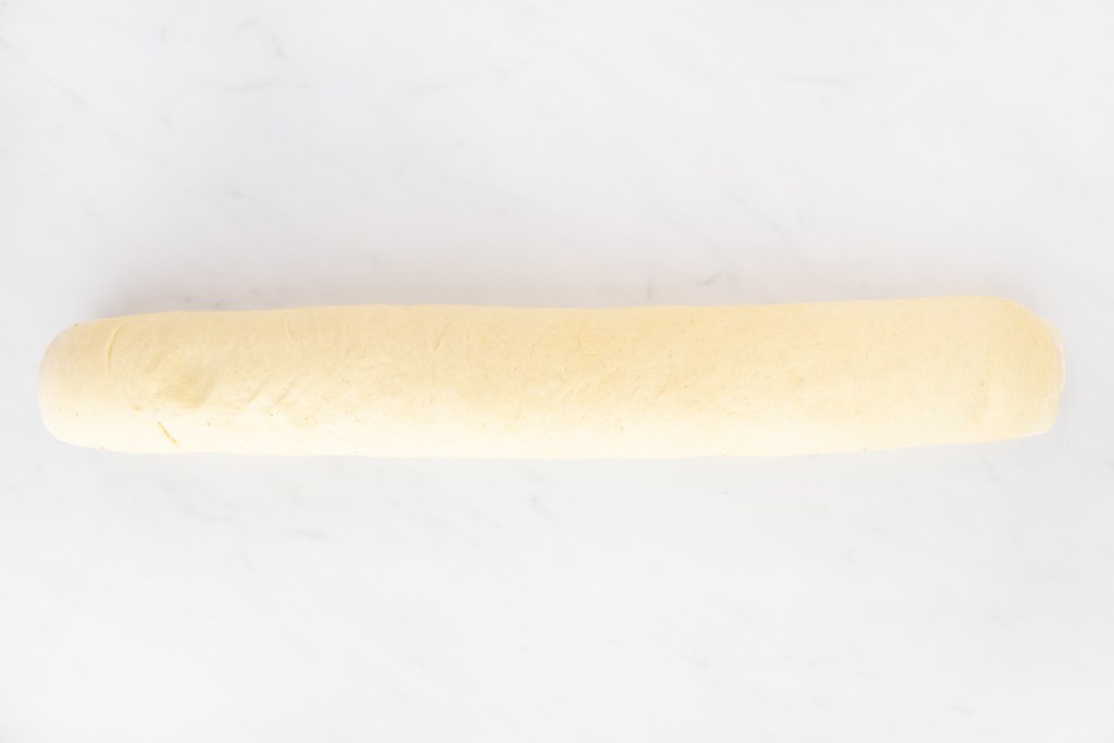 A long, rolled log of cinnamon roll dough on a white marble surface.