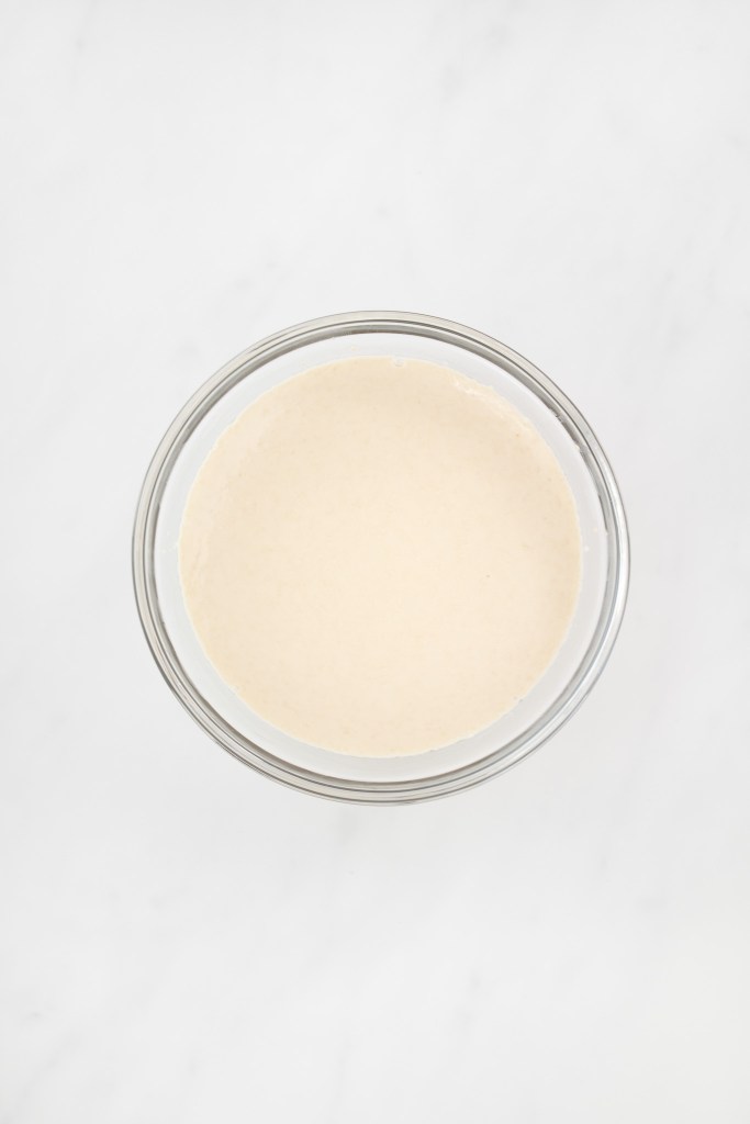A small glass bowl filled with a frothy proofed yeast mixture on a white marble surface.
