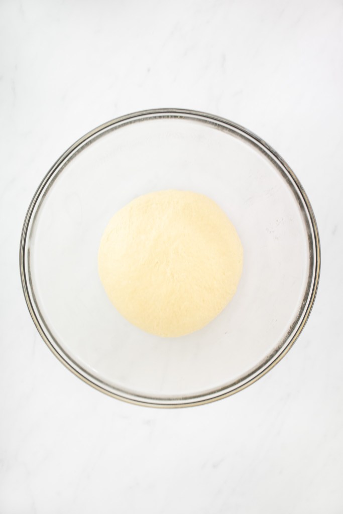 A smooth ball of un-risen cinnamon roll dough rests in the center of a clear glass bowl on a white marble surface.