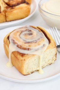 A cinnamon roll with icing drizzled on top sits on a white plate next to a fork, with more rolls and icing in the background.