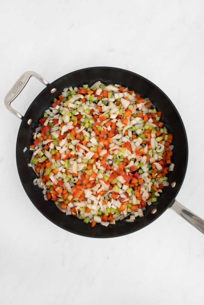 Chopped onions, celery, and red pepper are sautéing in a black skillet on a white marble surface.