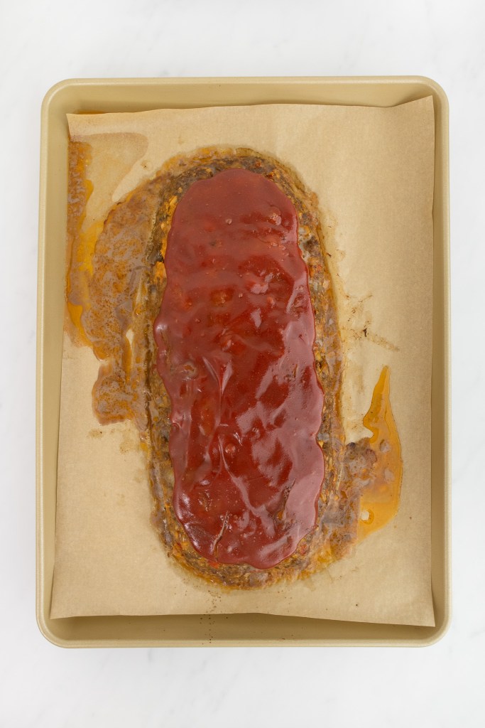 A fully baked meatloaf with red sauce on top sits on parchment paper in a rectangular sheet pan.