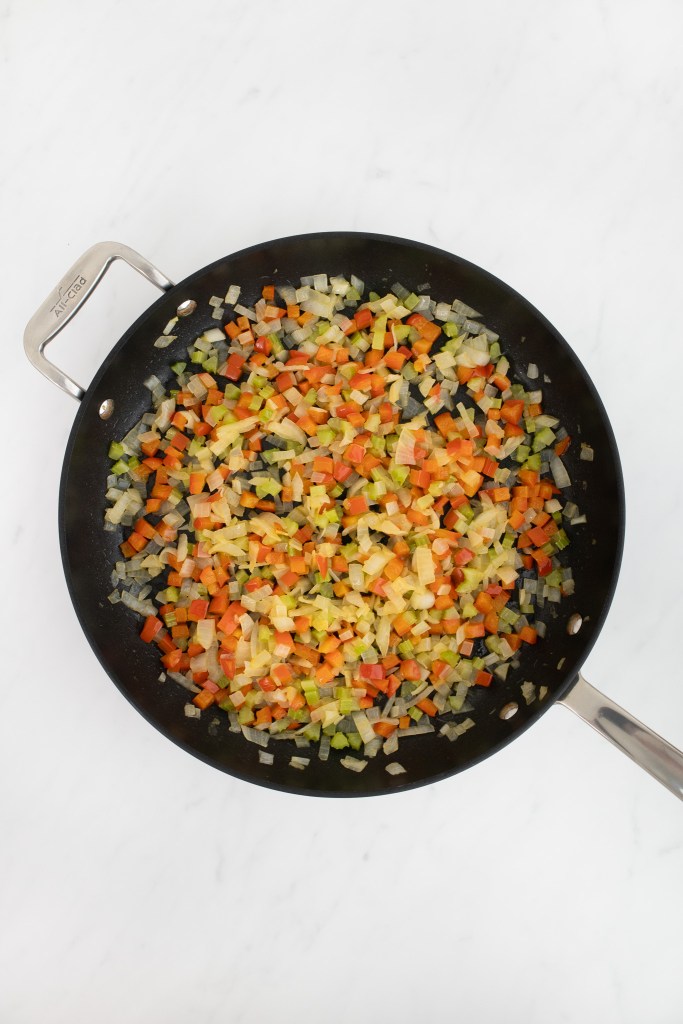 A frying pan on a white marble surface contains fully cooked and softened onions, celery and res pepper red.