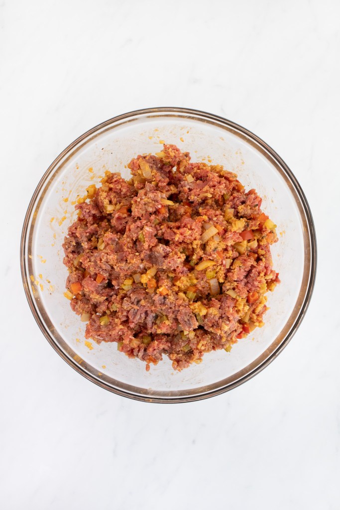A glass bowl filled with a fully combined meat loaf mixture sits on a white marble surface.