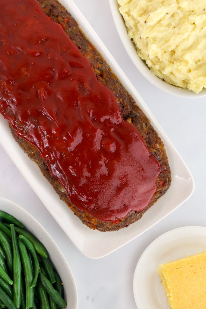 A meatloaf with red sauce is on a platter, next to bowls of green beans, mashed potatoes, and a square of cornbread.