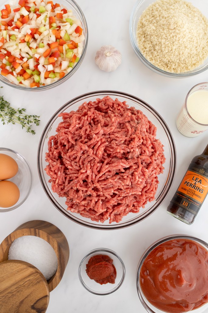 Ingredients for making meatloaf including: a bowl of ground beef, diced vegetables, breadcrumbs, eggs, tomato paste, ketchup, Worcestershire sauce, and seasonings, on a white marble surface.