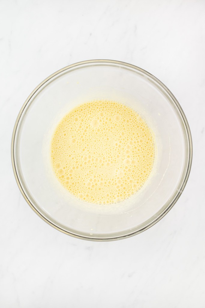 A glass bowl filled with a frothy, pale yellow liquid mixture sits on a white marble surface.