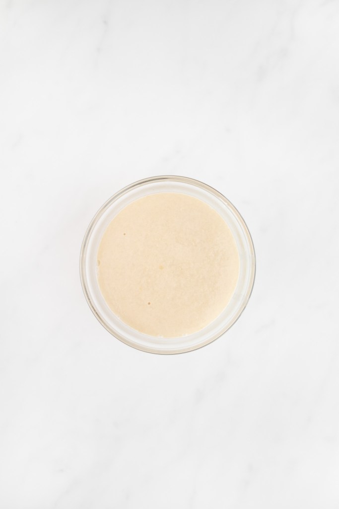 A small glass bowl filled with foamy, proofed yeast mixture sits on a white marble surface, viewed from above.