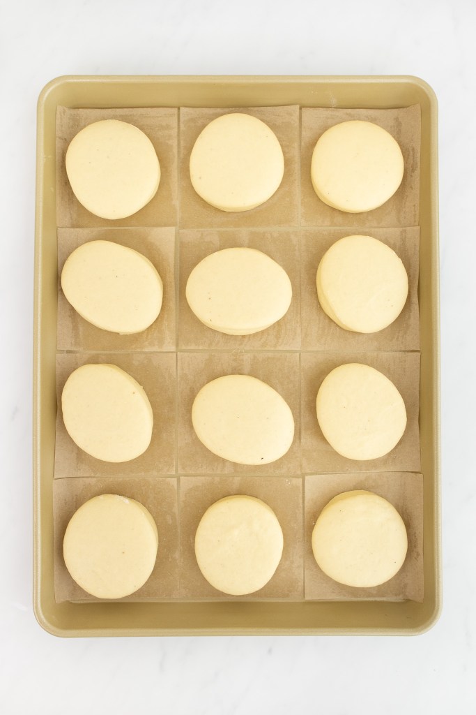 A baking tray with twelve evenly spaced, unbaked and risen donuts on parchment paper squares, viewed from above.