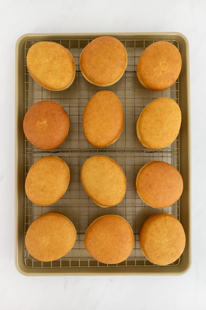 Twelve evenly spaced, round golden-brown donuts are on a wire rack set over a baking sheet, viewed from above on a white marble surface.