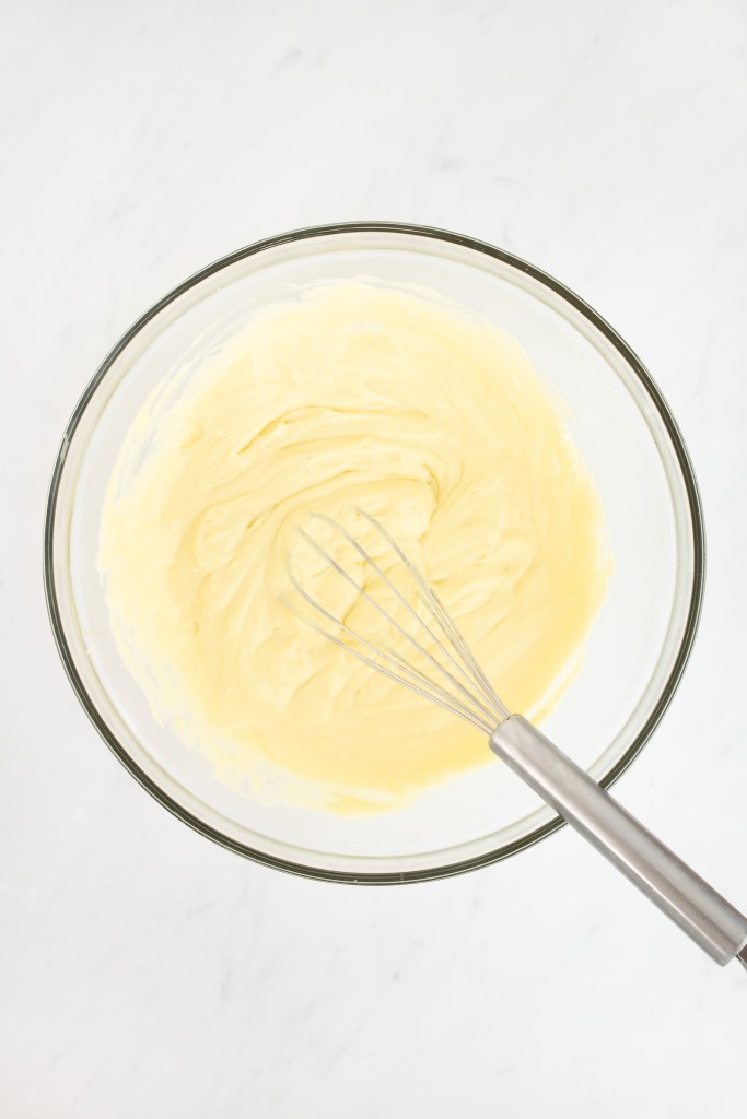 A metal whisk rests in a glass bowl filled with smooth pastry cream on a white marble surface.