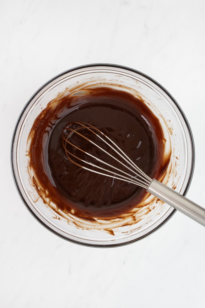 A metal whisk rests in a glass bowl filled with smooth, mixed chocolate ganache on a white marble surface.