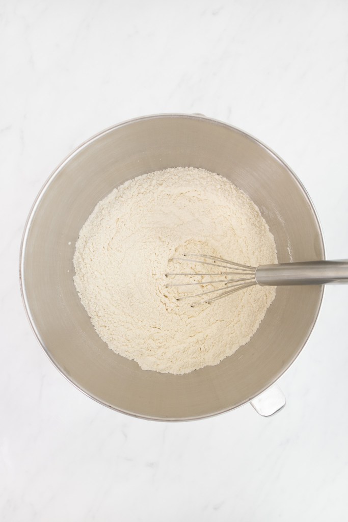 The bowl of an electric stand mixer with flour mixture for making donuts and a whisk inside, placed on a white marble surface.