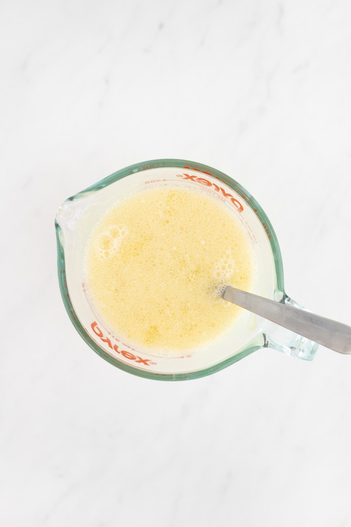 A glass measuring cup filled with liquid mixture for making donuts, on a white marble surface.