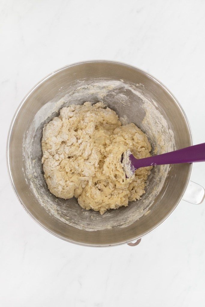 The bowl of an electric stand mixer with partially mixed dough and a purple spatula on a white marble surface.