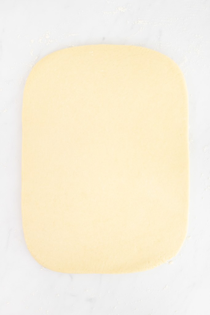 A rectangular sheet of rolled-out donut dough rests on a lightly floured white marble surface.