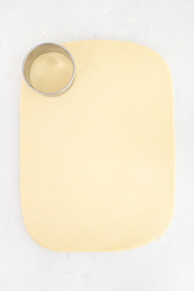 A metal biscuit cutter sits on rolled-out donut dough shaped in a rectangle on a lightly floured white marble surface.