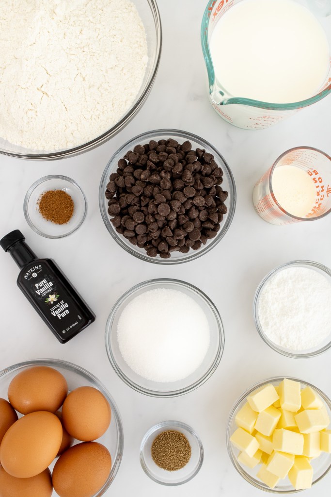 Ingredients for making Boston cream donuts including: flour, chocolate chips, sugar, eggs, butter, vanilla, milk, cream, nutmeg, and yeast on a white marble surface.