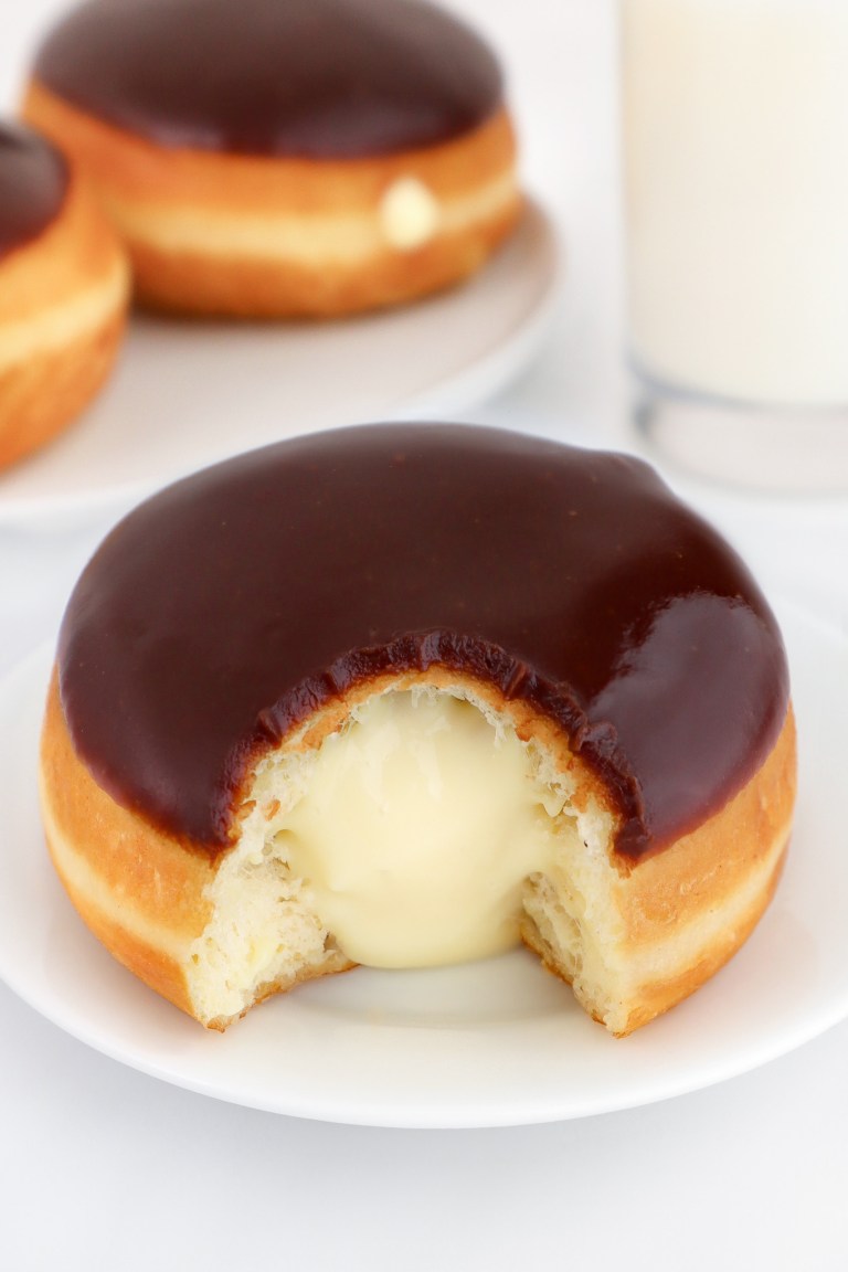 A Boston cream donut with a bite taken out, on a white plate; glass of milk and more donuts in the background.
