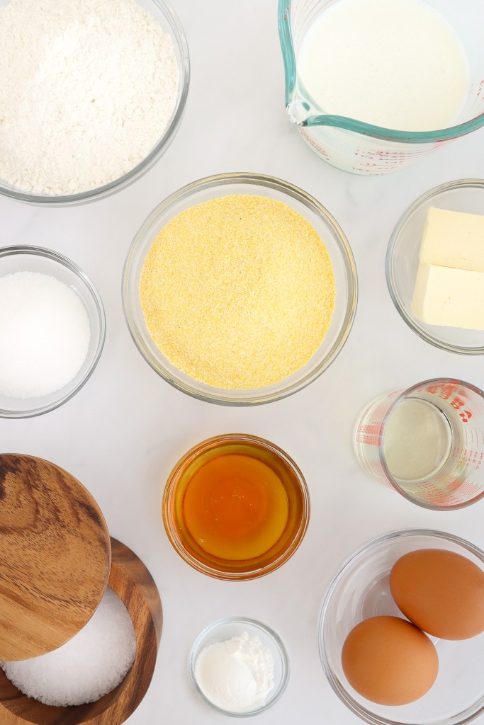 Ingredients for making cornbread muffins including: flour, cornmeal, eggs, butter, milk, sugar, honey, oil, salt, and baking powder, arranged on a white marble surface.