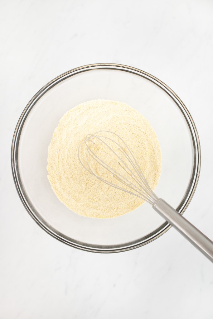 A metal whisk rests in a glass bowl filled with dry ingredients for making cornbread muffins, on a white marble surface.