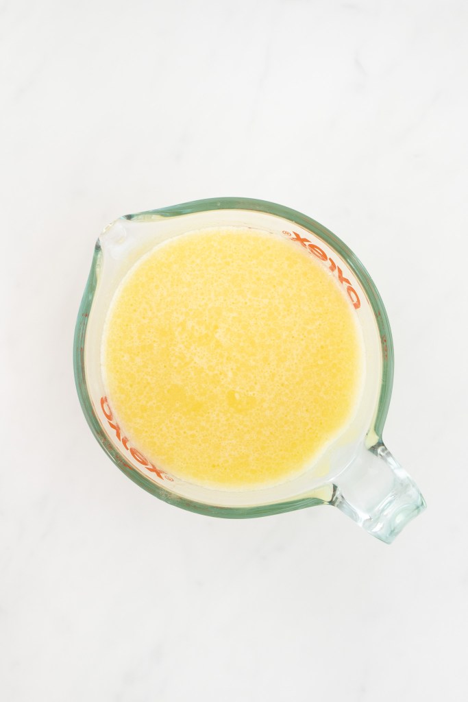 A glass measuring cup filled with a wet ingredients for cornbread muffins, viewed from above, on a white marble surface.