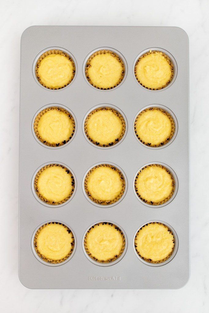A muffin tin lined with paper cups holds twelve portions of unbaked cornbread muffins on a white marble surface.