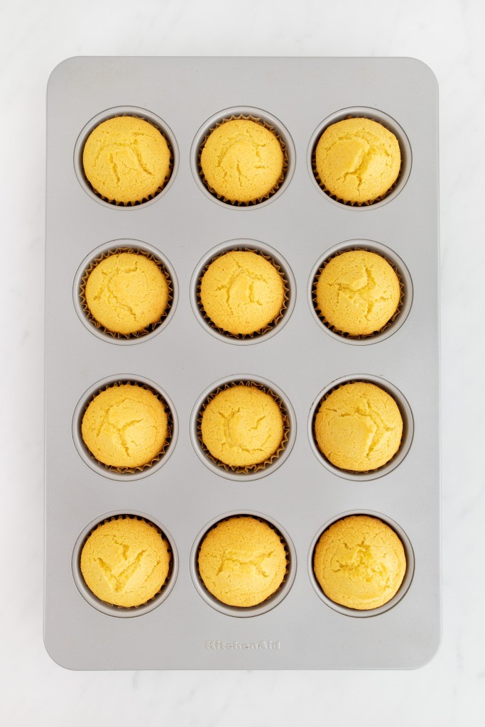 A metal muffin tin holds twelve evenly baked cornbread muffins in paper liners on a white marble surface.