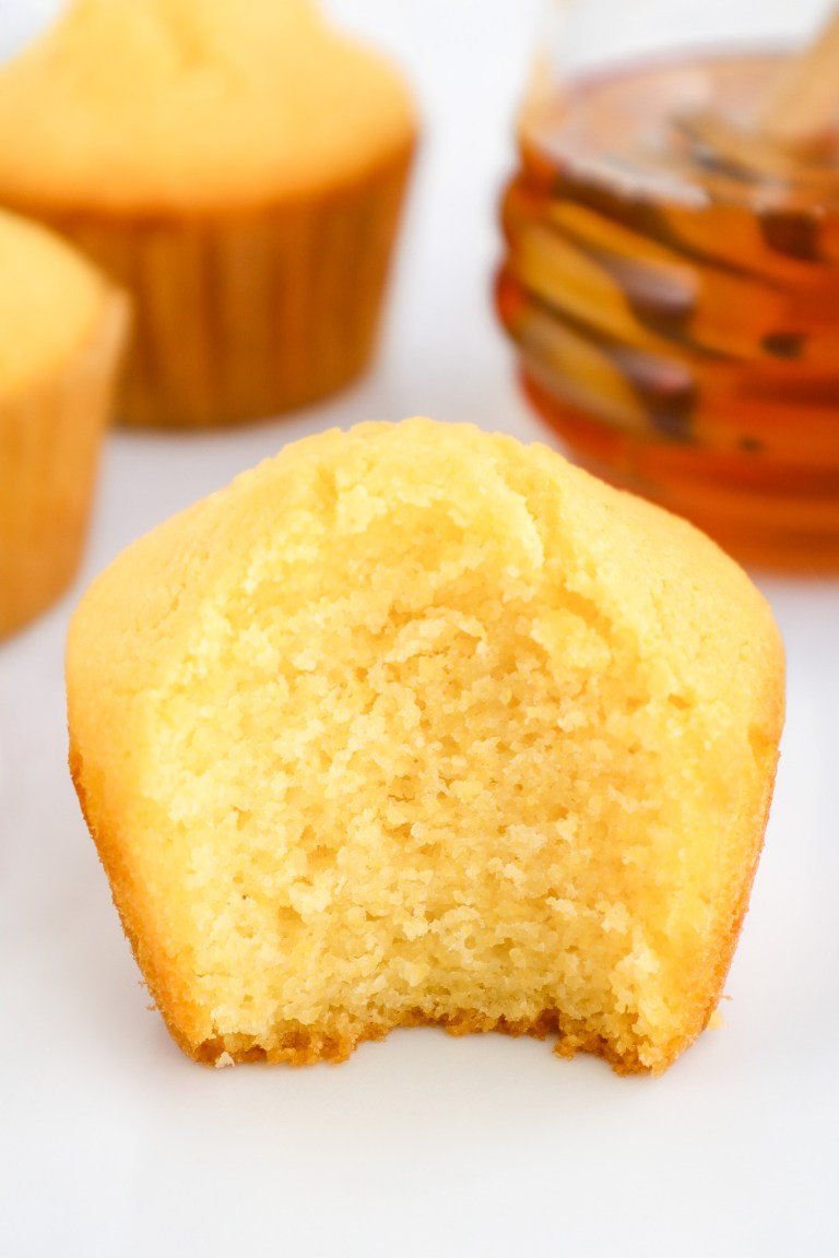 A cornbread muffin with a large bite taken out, placed in front of a jar of honey and other muffins on a white marble surface.