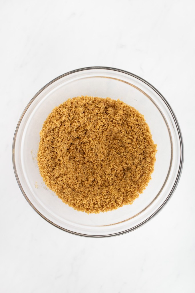 A glass bowl filled with graham cracker crust mixture sits on a white marble surface.